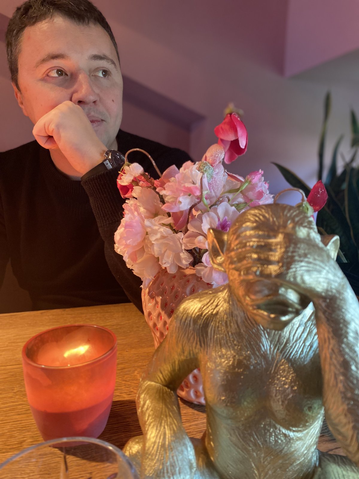 A man with his hand to his chin at a table with flowers and a gold-coloured monkey figure.