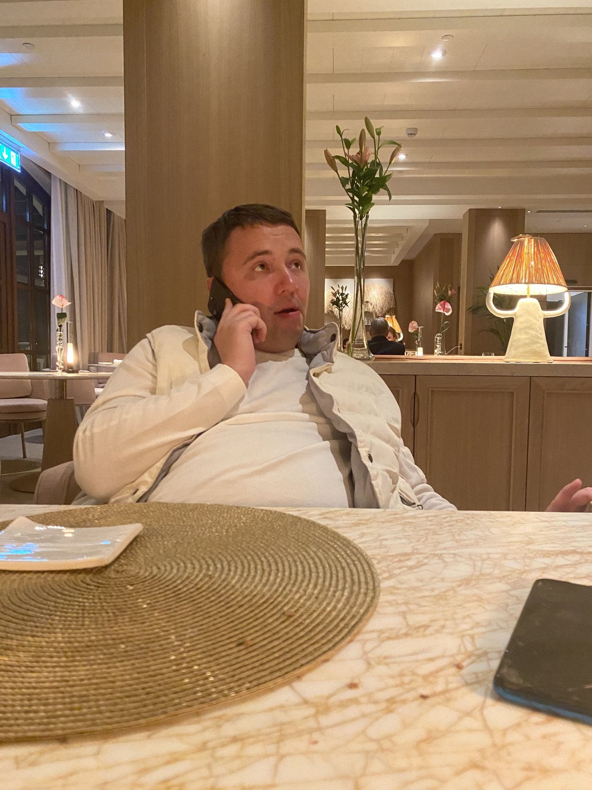 A man in a light jacket on the phone at a restaurant table.