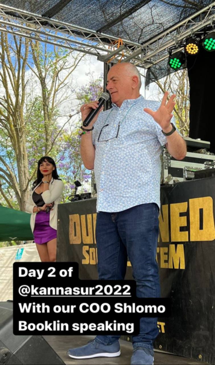 A man with a microphone on an outdoor stage in front of a banner. Overlaid text reads: Day 2 of at kannasur2022, With our COO Shlomo Booklin speaking.