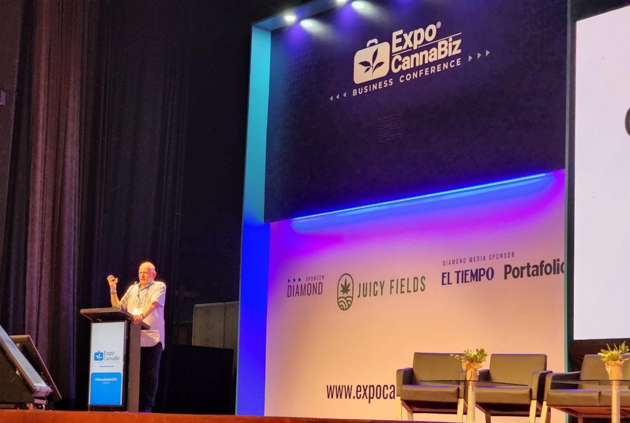 A speaker at a lectern at ExpoCannaBiz. Behind him a large wall with the organiser's logo and sponsor logos, among them Juicy Fields and El Tiempo.