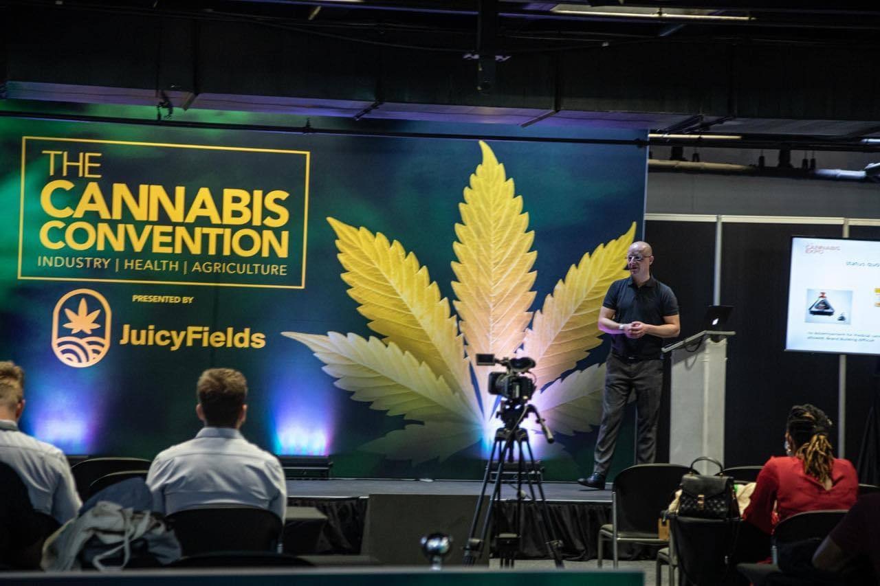 A trade fair stage with a large screen. The screen reads THE CANNABIS CONVENTION, INDUSTRY HEALTH AGRICULTURE, PRESENTED BY JuicyFields, beside a large yellow cannabis leaf. At the lectern on the right stands a man in glasses and a dark polo shirt. An audience sits in front of the stage and a cameraman films.