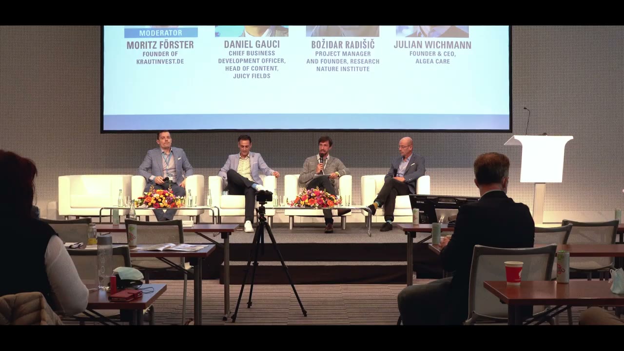A panel on stage in front of an audience. The screen behind them carries the names and titles: MODERATOR Moritz Förster, Founder of krautinvest.de, and Daniel Gauci, Chief Business Development Officer, Head of Content, Juicy Fields.