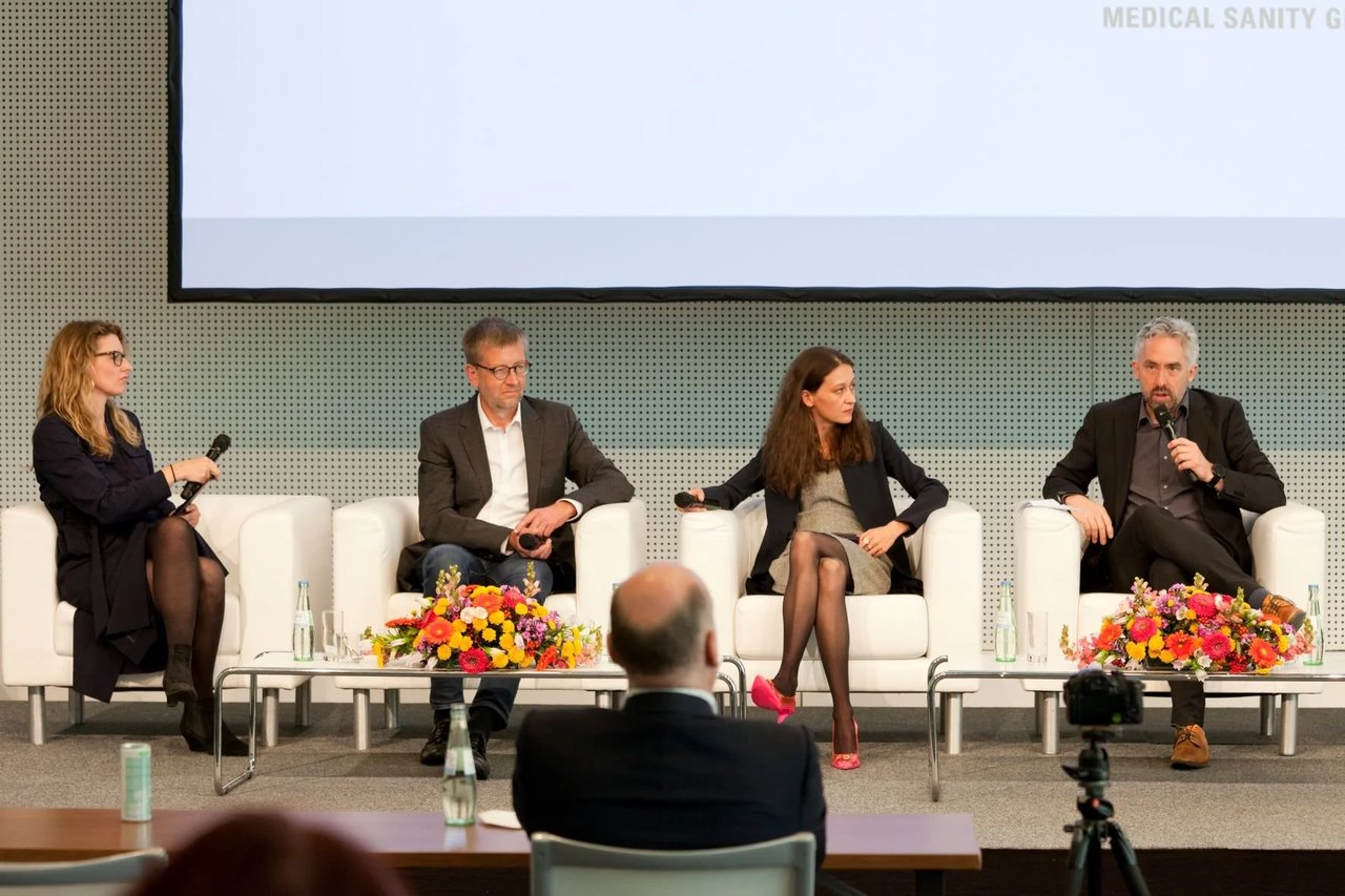 Four people in white armchairs on a stage, with flower arrangements in front of them and a projection screen behind. On the wall to the right the words MEDICAL SANITY GROUP are visible.