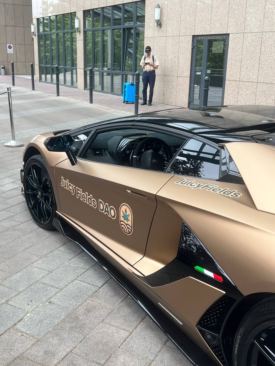 A gold-coloured Lamborghini parked on a paved street outside a glass facade. On the door the text Juicy Fields DAO with a logo, and behind the car a person stands at the entrance.