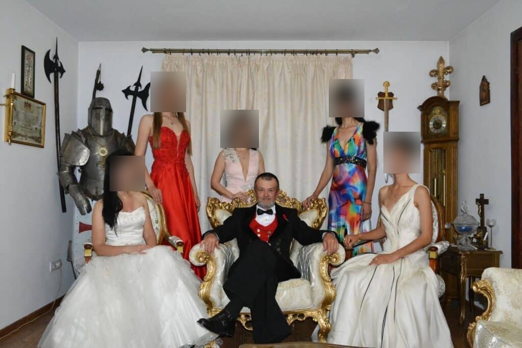 A man in a tuxedo seated in a gilded throne chair, surrounded by five young women in evening and wedding gowns, with a suit of armour and halberds in the room. The women's faces are blurred.