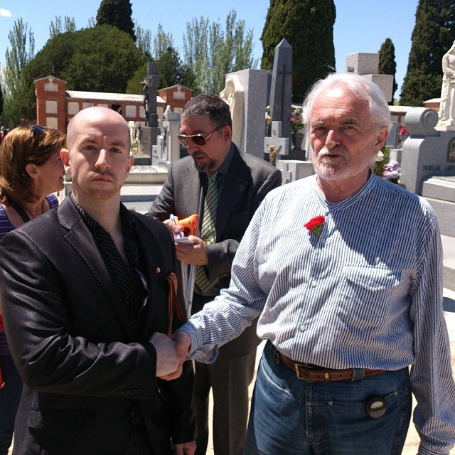 Two men shake hands in a sunny cemetery. On the left a bald man in a dark jacket, on the right an older white-haired man in a striped shirt with a red carnation.