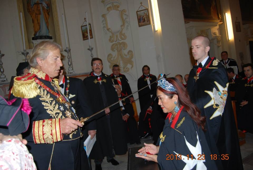 A man in a gold-embroidered uniform holds a sword over a kneeling woman in an order's mantle and tiara, in a church. Behind them stand a bald young man in an order's mantle and several knights.