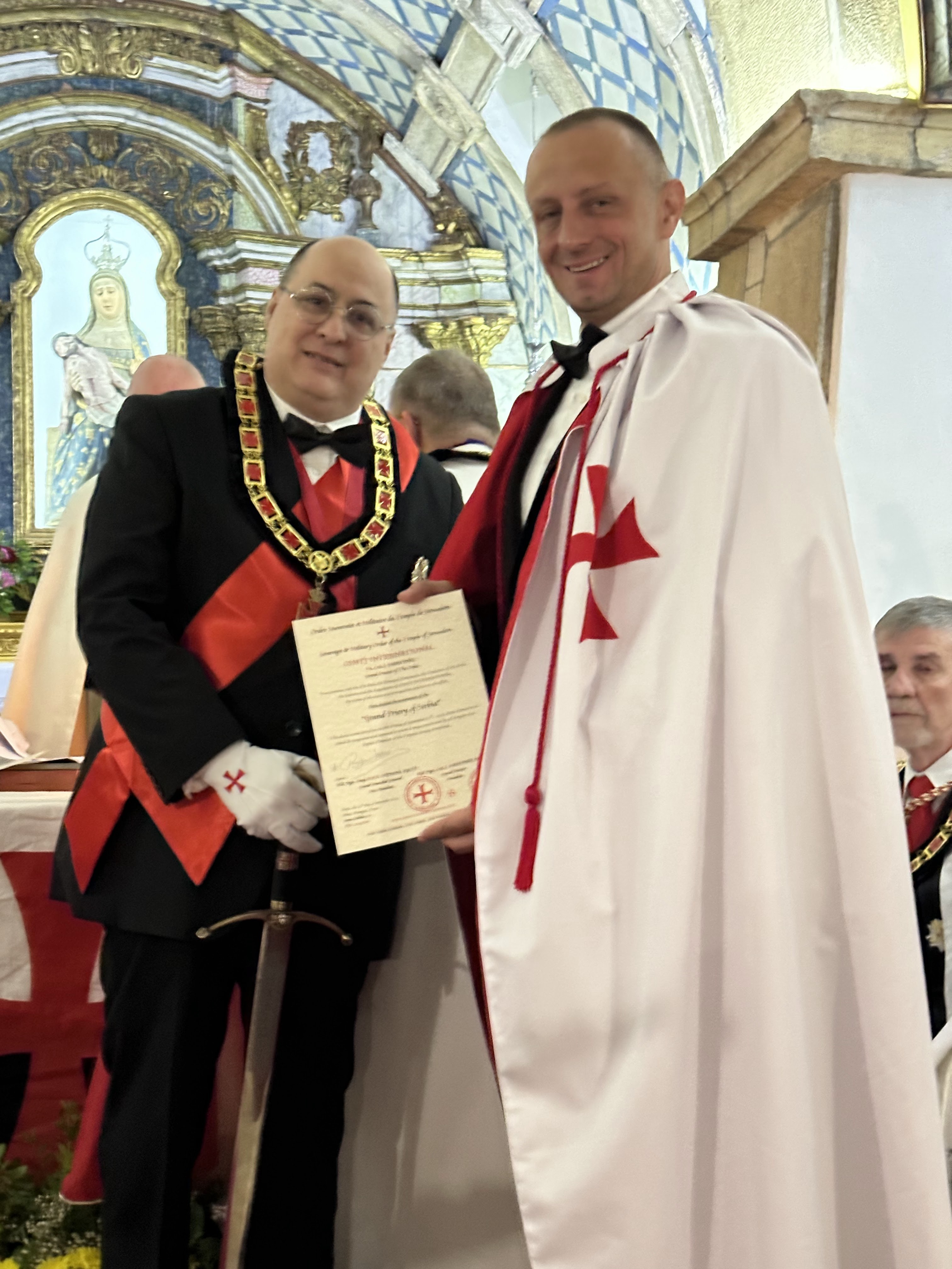 A man in a white mantle with a red Templar cross holds up a certificate together with a man wearing an order chain and a sword, in a church.