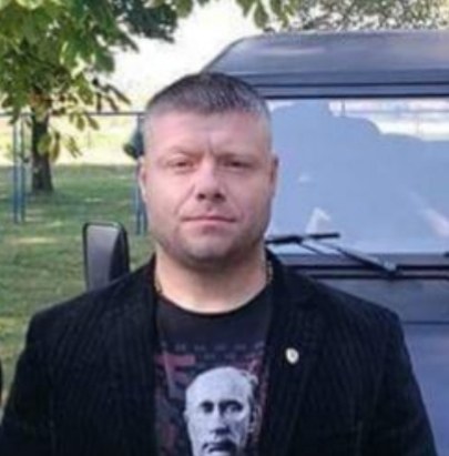 A man with short hair in a dark jacket in front of a black off-road vehicle. Under the jacket a black T-shirt printed with Vladimir Putin's face.