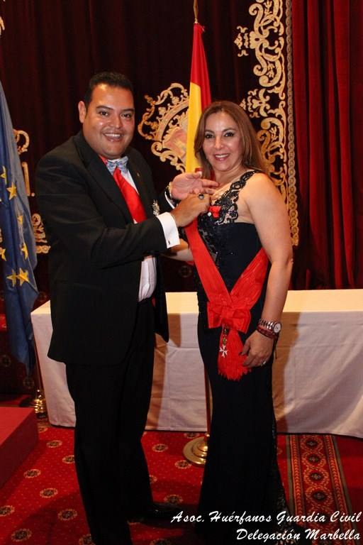 A man in a tuxedo places a red sash with a cross on a woman in a black evening gown in front of a Spanish flag and an EU flag.