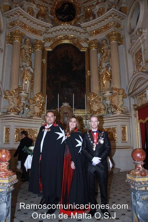 Three people in black order mantles with white eight-pointed crosses stand in front of a gilded baroque altar.